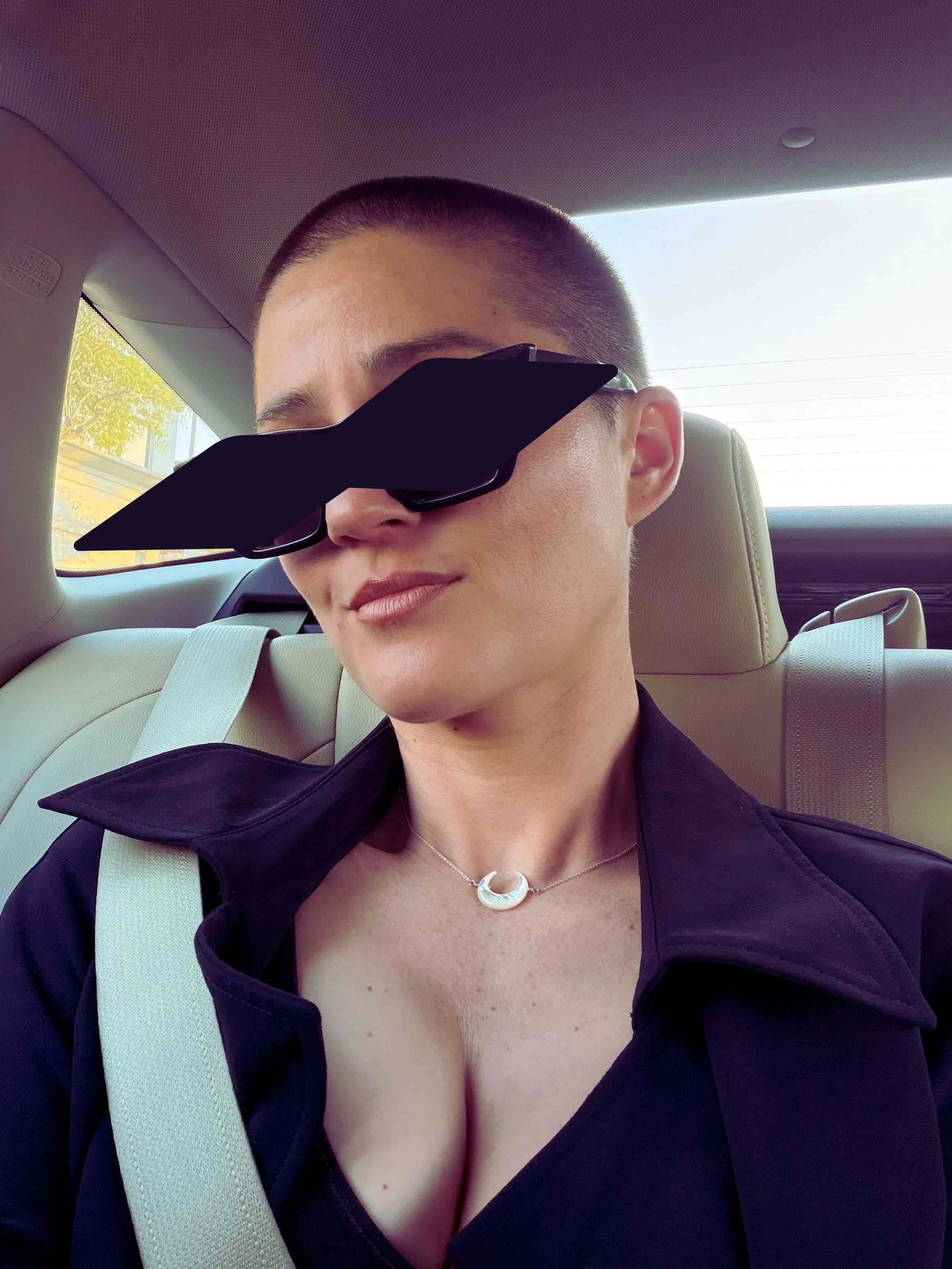 Close-up portrait of a person with a shaved head wearing dark sunglasses and a necklace, seated in a car.